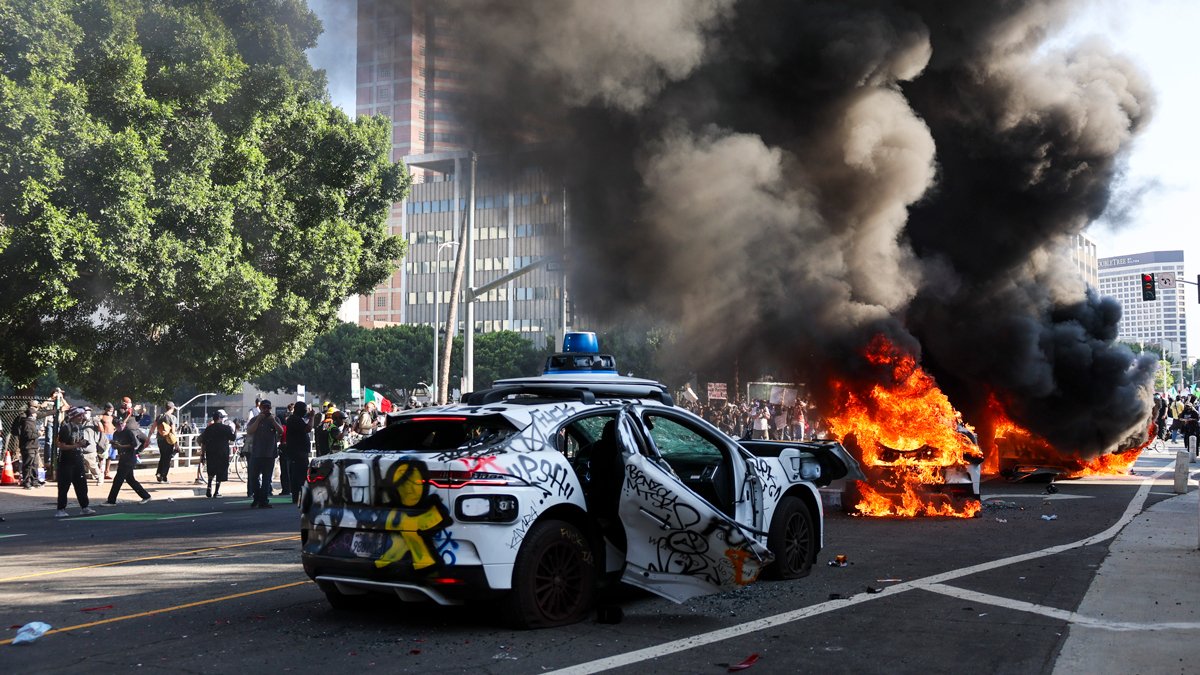 Горящие автомобили на фоне протестующих, Лос-Анджелес, 8 июня 2025 года. Фото: Allison Dinner / EPA